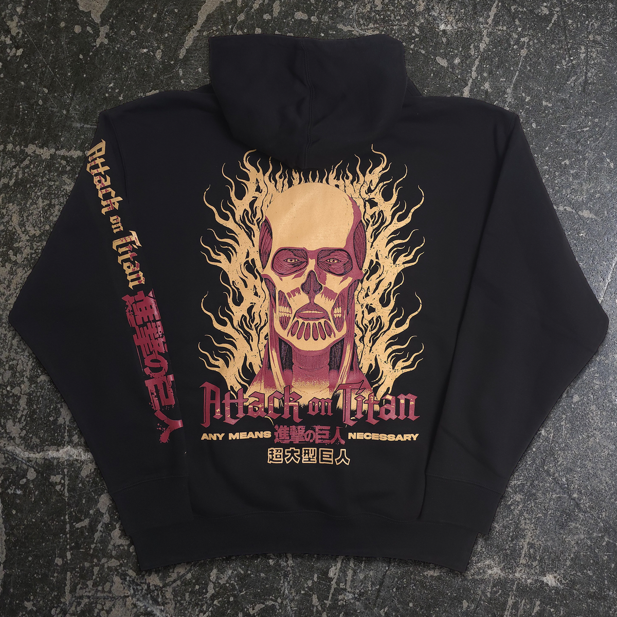 A black hoodie laid flat on a dark textured surface, featuring a large Attack on Titan graphic on the back with a skull surrounded by flames in beige, pink, and red, plus Japanese text printed down the sleeve.