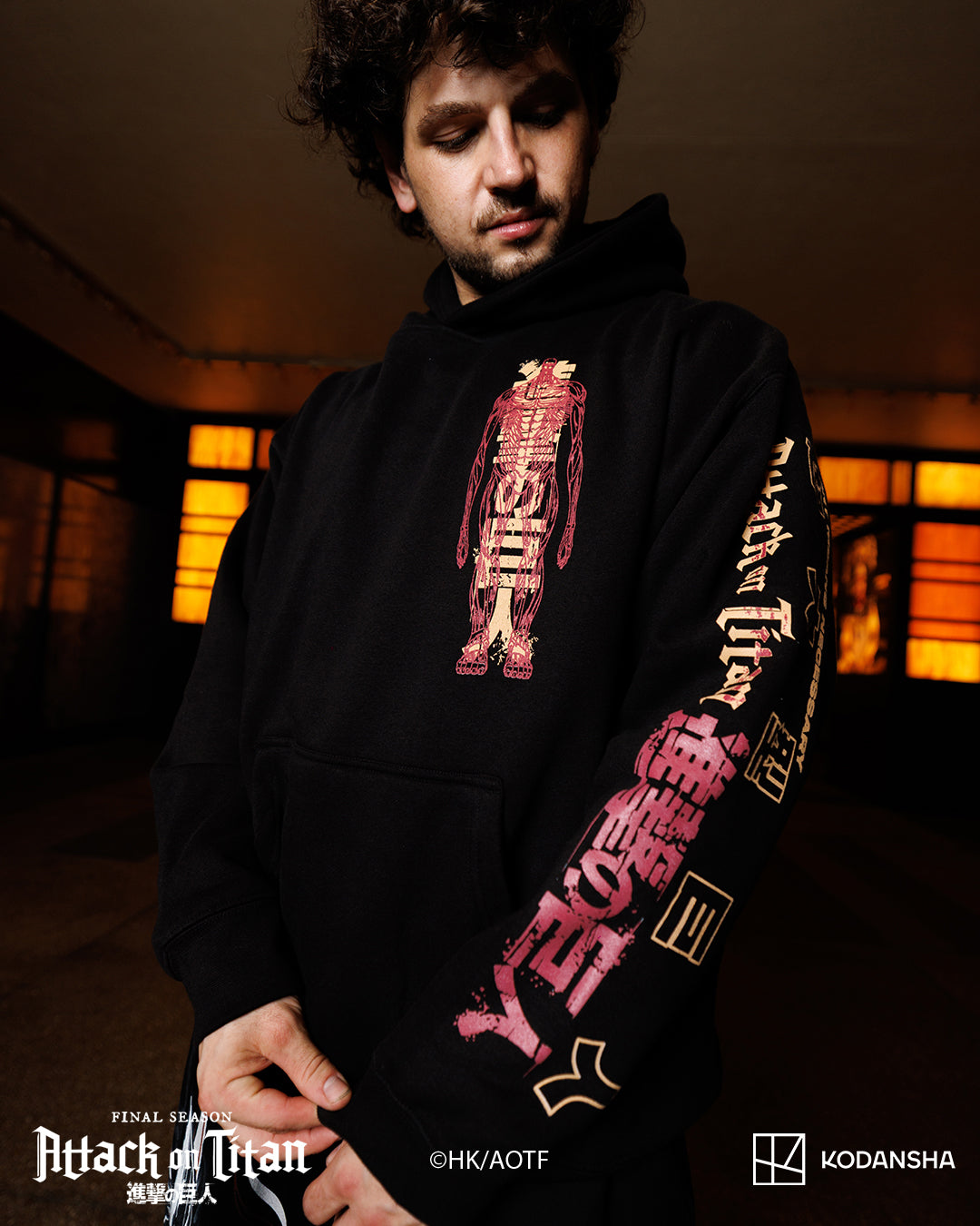 A man wearing a black Attack on Titan themed hoodie with red and gold Japanese text graphics on the front and sleeves, set in a dimly lit indoor environment.