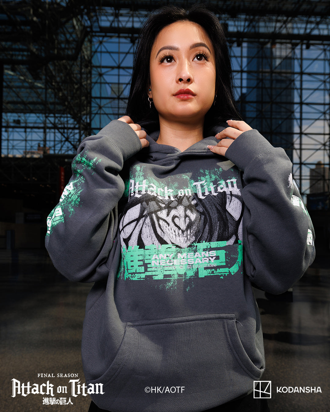 A woman wearing a dark gray Attack on Titan Final Season hoodie with a wolf graphic and green text, posed in front of a dark industrial background. The image includes Attack on Titan Final Season and Kodansha logos at the bottom.