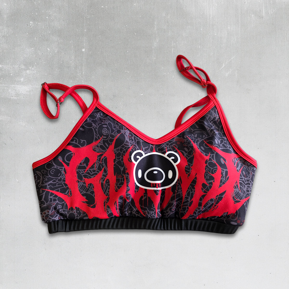 A women's sports bra with red and black graphic details, featuring a central cartoon bear face logo and thin adjustable straps, laid flat on a textured gray background. gloomy bear