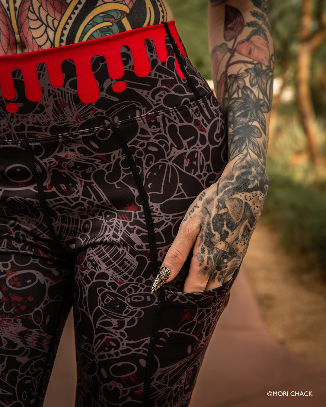 Close-up of a person wearing black leggings with a gray line-art tattoo-style print and a bright red waistband with dripping red trim. The person's tattooed hand and torso are visible against an outdoor blurred background. Gloomy Bear fitness