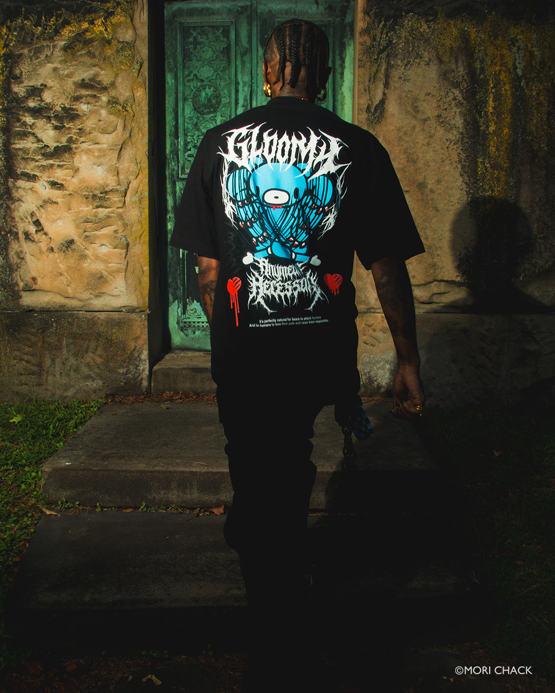 A man viewed from behind wearing a black t-shirt with a large blue, white, and red graphic on the back, standing in front of a dark, mossy green door in a dimly lit stone building.