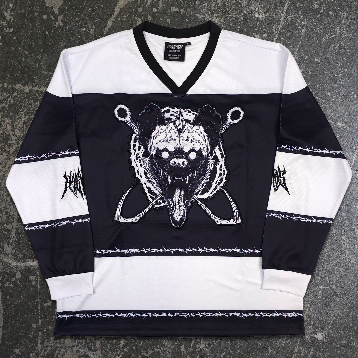 A black and white long-sleeve hockey-style jersey laid flat on a dark textured surface. The jersey features a black V-neck collar, a large black-and-white bear skull graphic with crossed tools on the chest, white sleeve panels, barbed wire stripe details, and black graphic text on the sleeves. any means necessary shawn coss goon squad hockey jersey