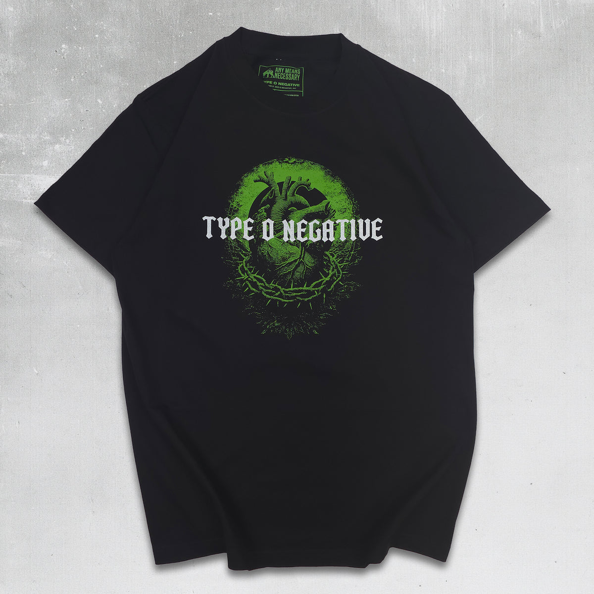 A black short-sleeve graphic T-shirt centered on a dark green textured background with thorny branches framing the edges. The shirt features a bright green circular graphic with an anatomical heart and the words 'TYPE O NEGATIVE' in white across the chest.