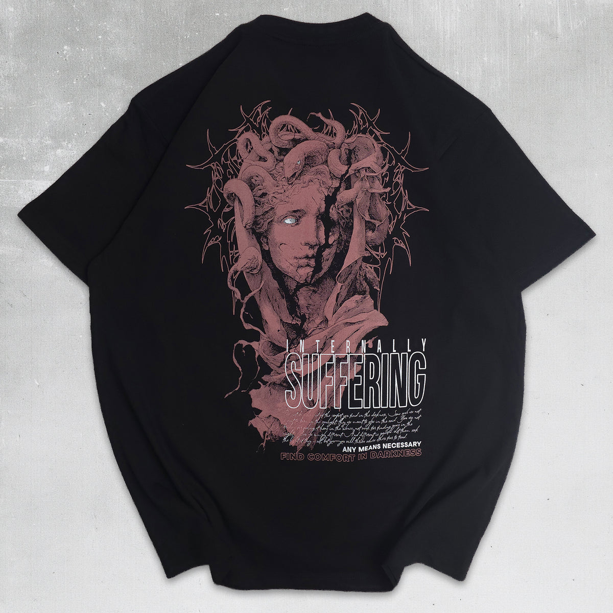 A black short-sleeve graphic t-shirt laid flat on a textured gray background, featuring a large back print of Medusa with snakes for hair in a dusty rose color, with the word 'SUFFERING' in a distressed white font.