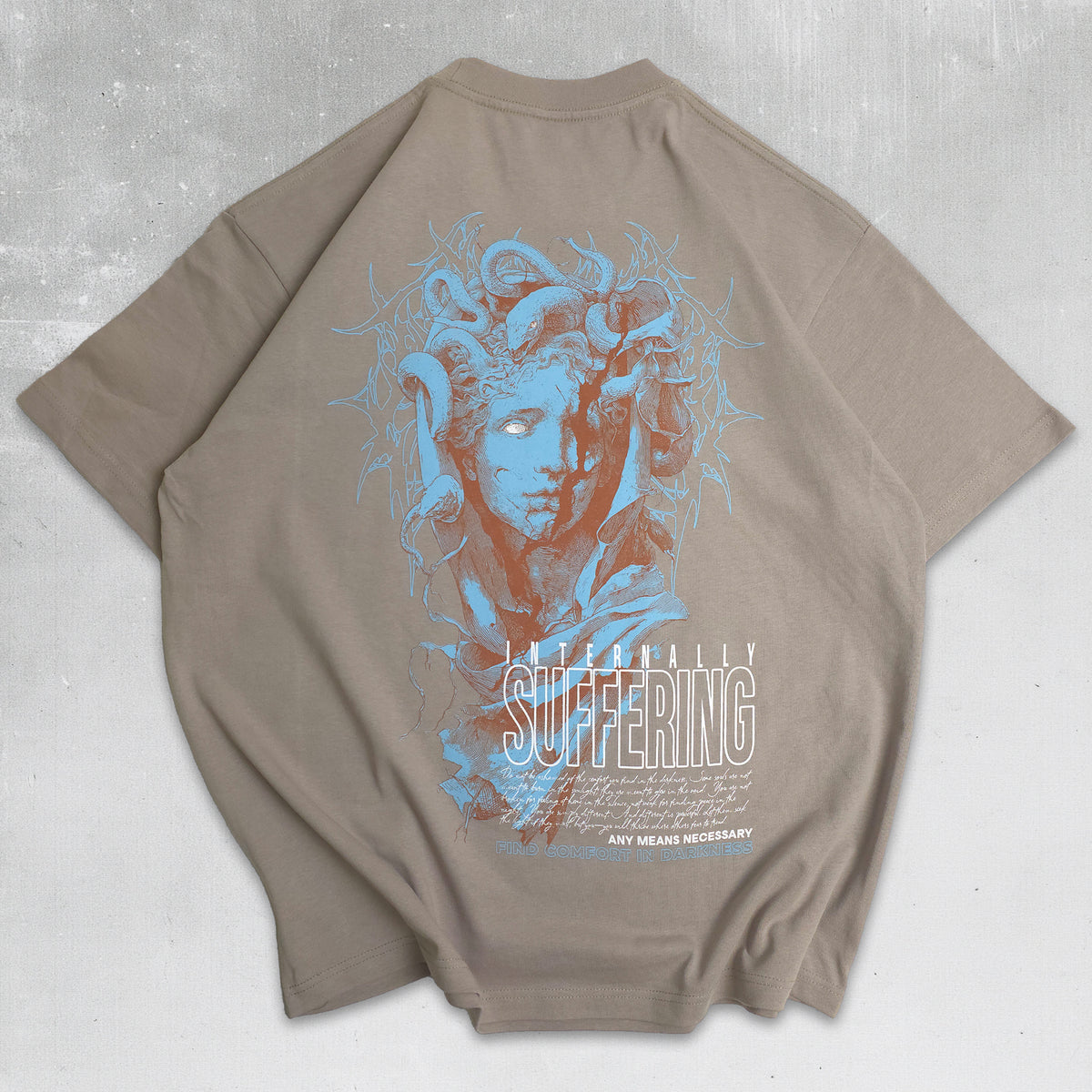 A taupe-beige short-sleeve t-shirt laid flat on a textured gray background, featuring a large graphic print of Medusa in shades of blue and orange-brown, with the word 'SUFFERING' printed below.
