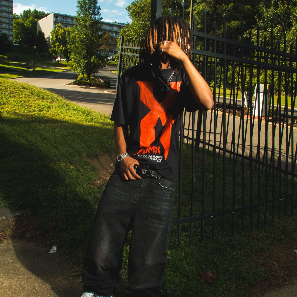 A person standing outdoors on a sidewalk next to a black metal fence, wearing a black graphic t-shirt with a large orange and white design, dark baggy jeans, and white sneakers. DMX