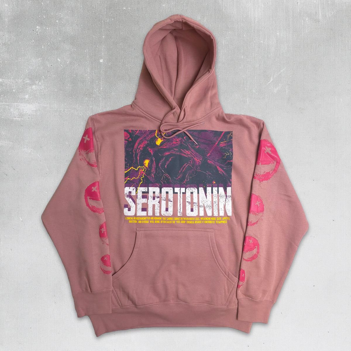 A dusty rose pink pullover hoodie with a graphic print on the front featuring the word 'SEROTONIN' in white, a dark abstract artwork, and pink circular patterns on the sleeves, laid flat on a textured gray background.