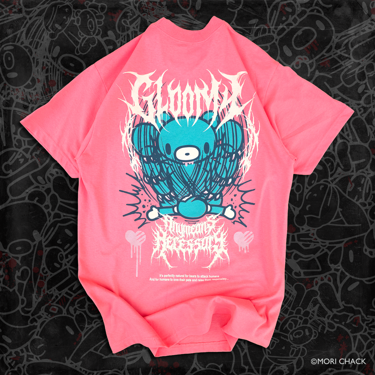 A bright pink short-sleeve t-shirt featuring a central graphic of a blue cartoon bear with the word 'GLOOMY' above and 'Any Means Necessary' below in a stylized font, laid flat on a black patterned background.