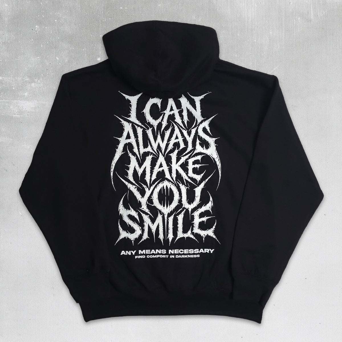 A black pullover hoodie laid flat on a textured gray background, featuring a large white spiky gothic text graphic on the back that reads 'I CAN ALWAYS MAKE YOU SMILE' with smaller text below reading 'ANY MEANS NECESSARY' and 'AND UNCONDITIONAL UNDERMINING'.