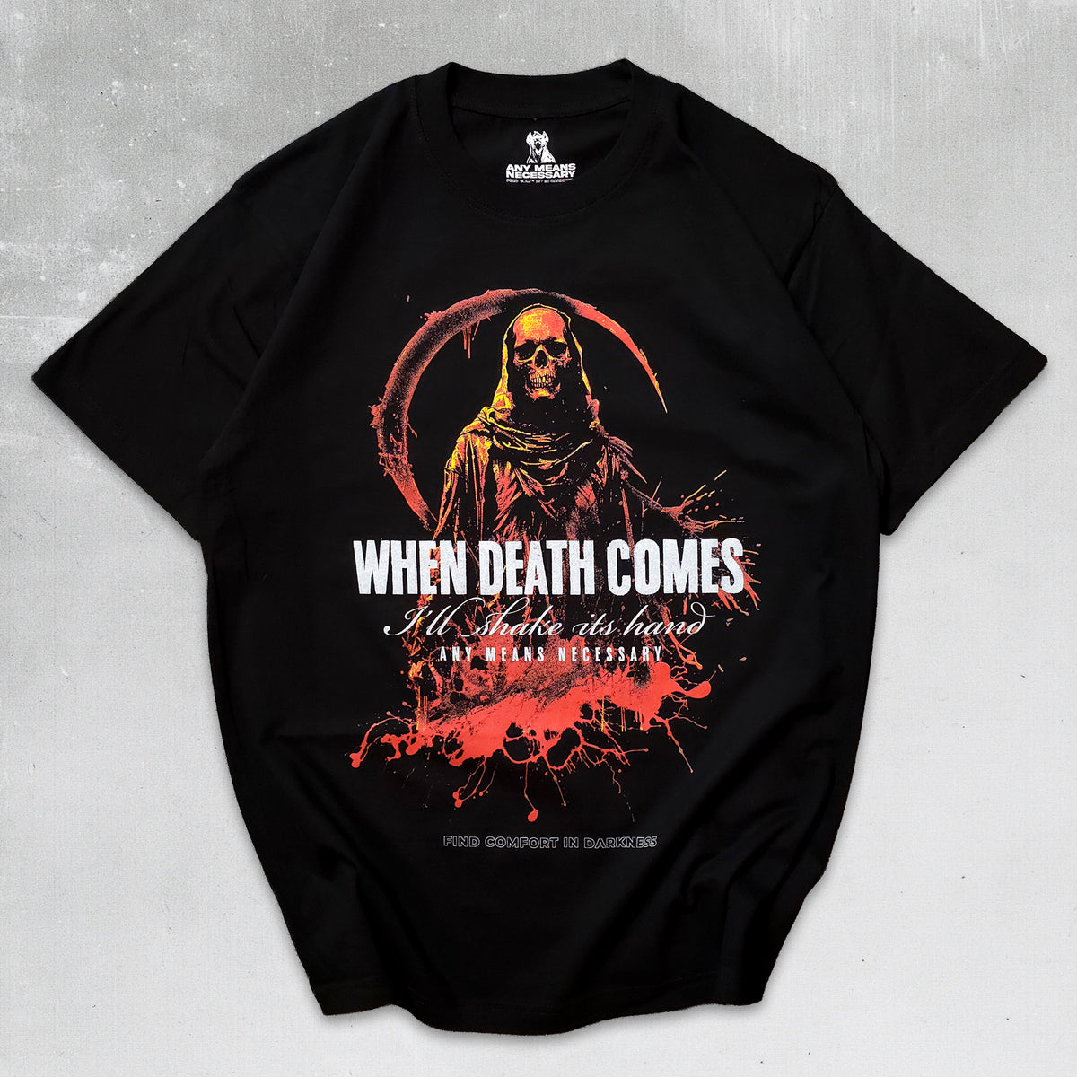 A black short-sleeve graphic t-shirt laid flat on a textured gray background. The shirt features a grim reaper design with a skull and scythe in red and orange tones, with the text 'WHEN DEATH COMES I'll Make it hers' and 'THE DEAD BIRTHDAY' printed in white and red.