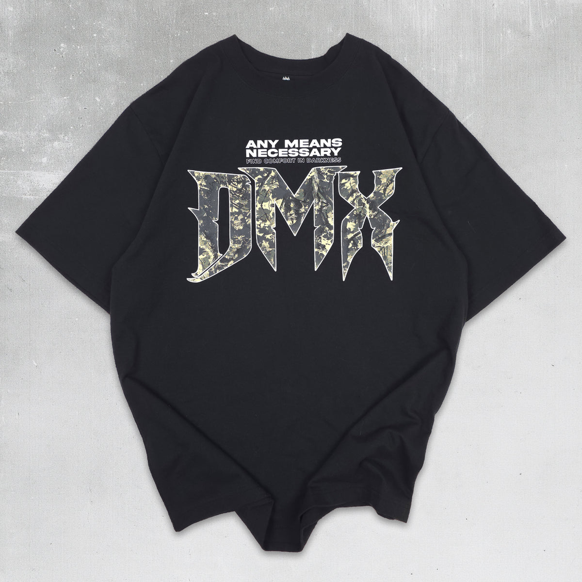 A black oversized short-sleeve t-shirt with a graphic print on the front. The print features the text 'ANY MEANS NECESSARY' in white above a large stylized 'DMX' logo filled with a green and beige camouflage pattern, laid flat on a textured gray background.