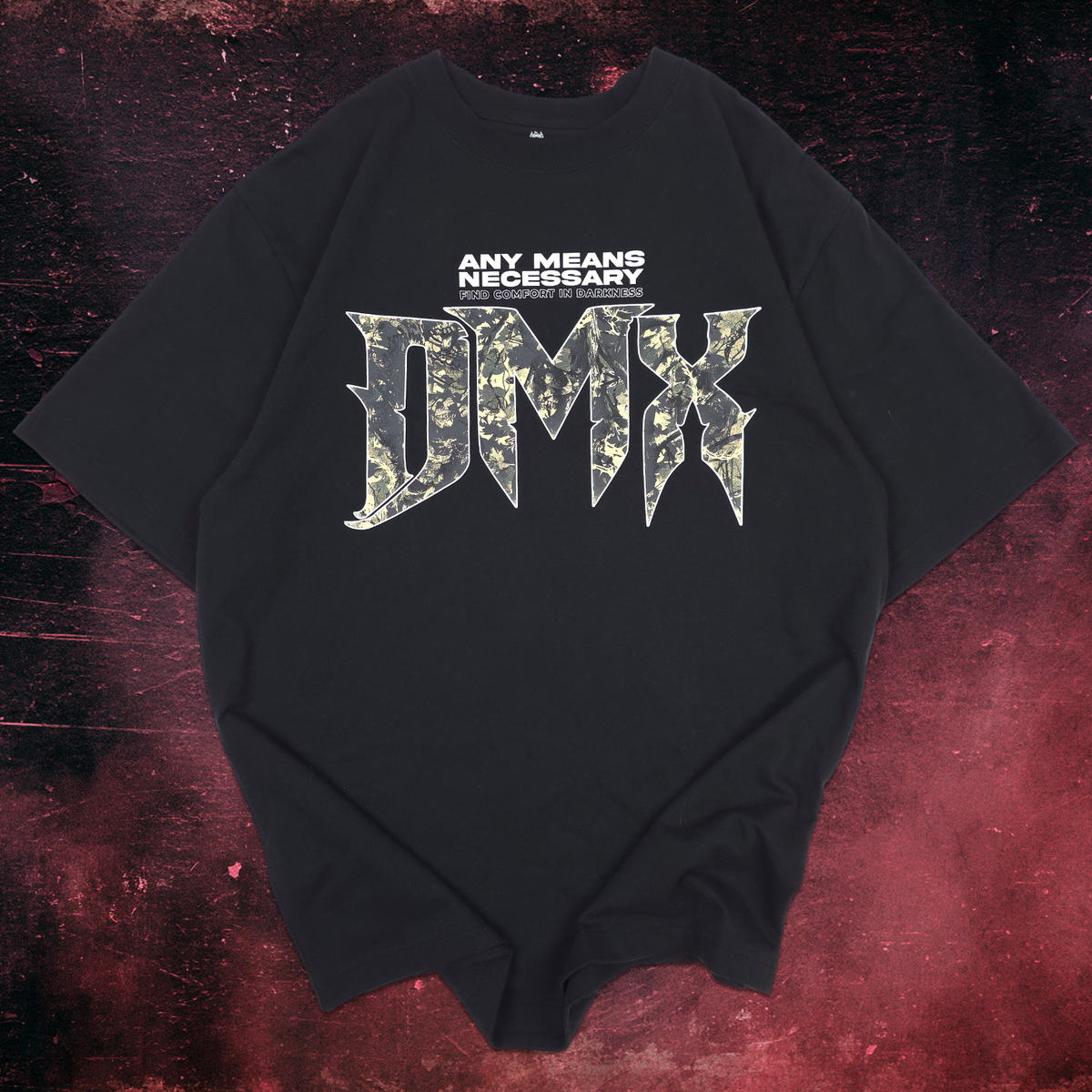 A black oversized t-shirt with a graphic on the front reading 'ANY MEANS NECESSARY DMX' in white and camouflage lettering, displayed flat against a dark red and black textured background.